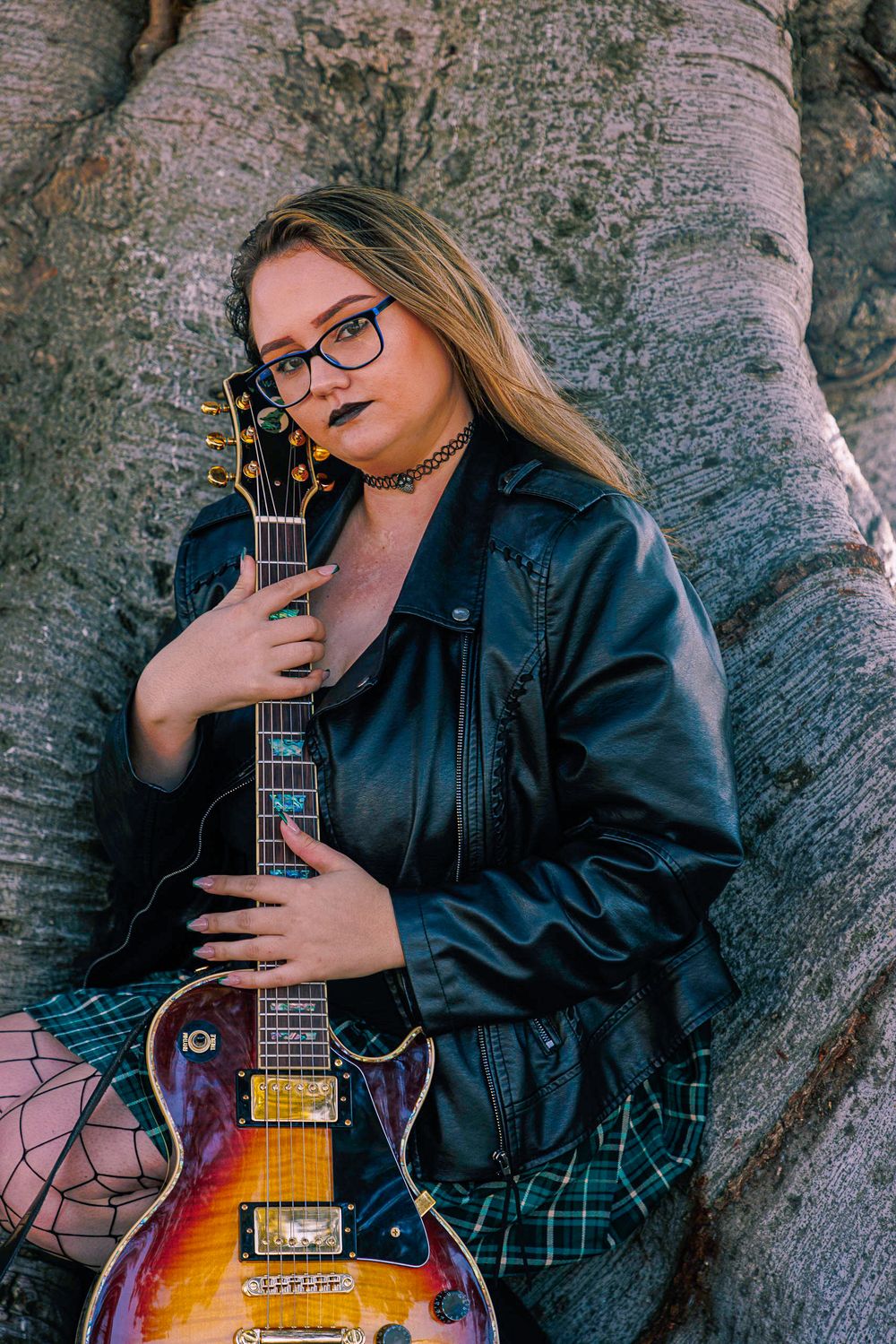 Young woman holding a sunburst electric guitar while leaning against a large textured tree trunk.