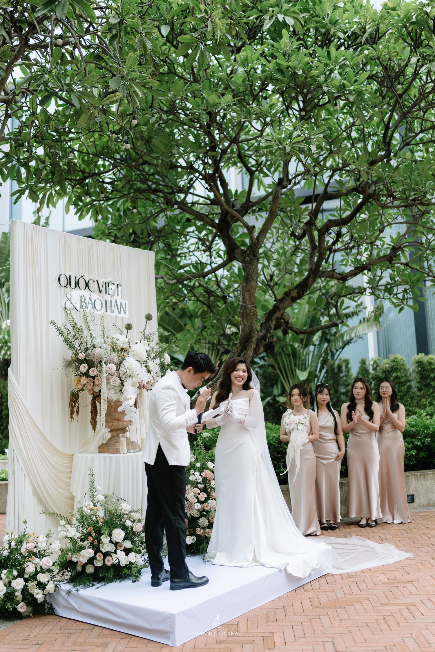 Couple exchanges rings at outdoor wedding ceremony with white floral backdrop and bridesmaids in champagne dresses.