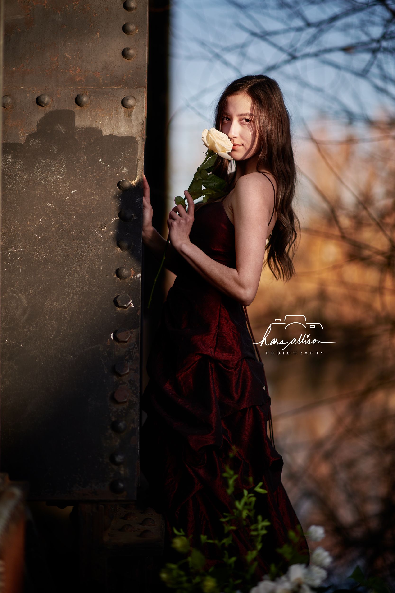 A moody portrait against industrial metal shows someone in a dark gown holding a delicate white rose at sunset.