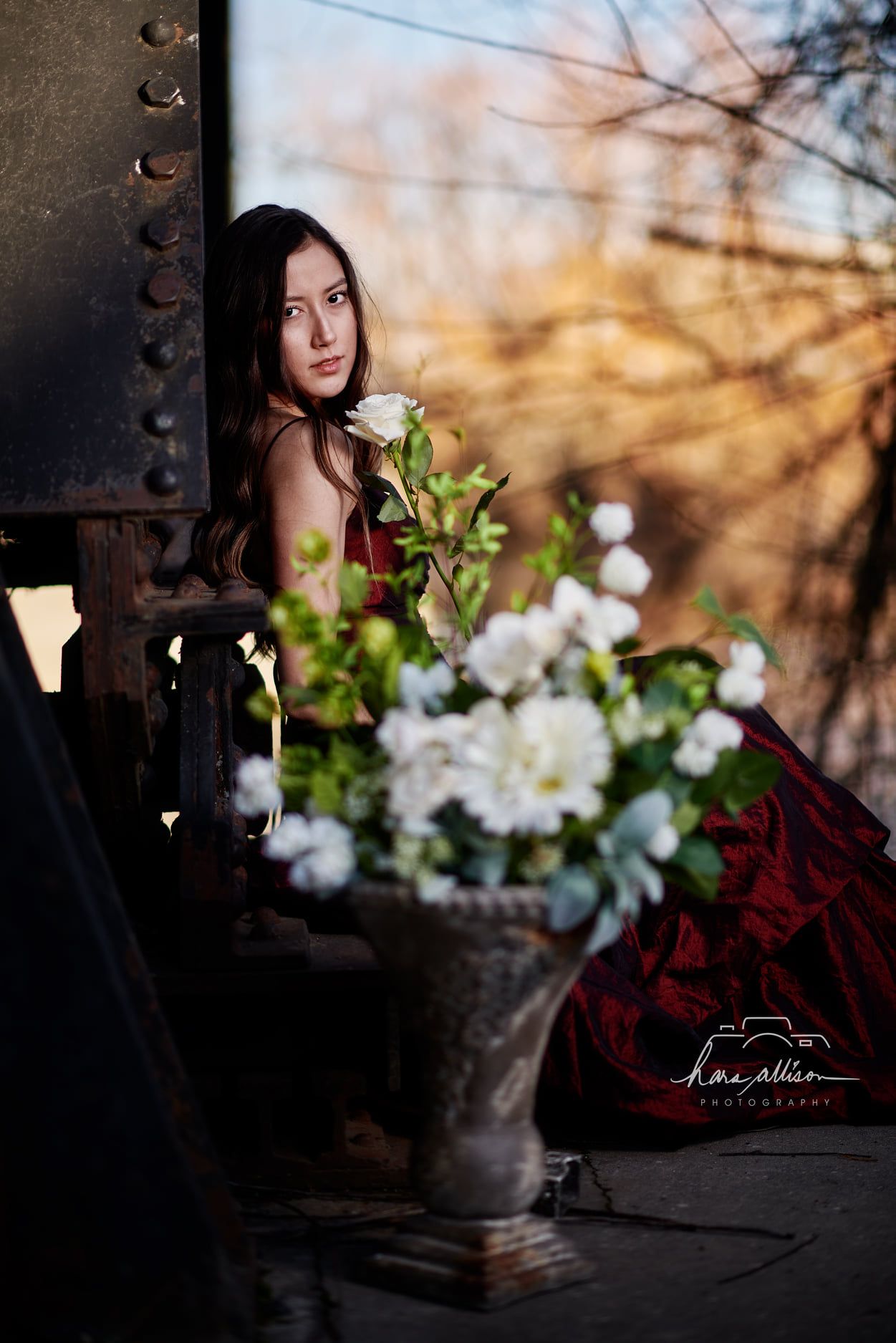 A romantic portrait with burgundy dress and white floral arrangement against industrial bridge elements at sunset.