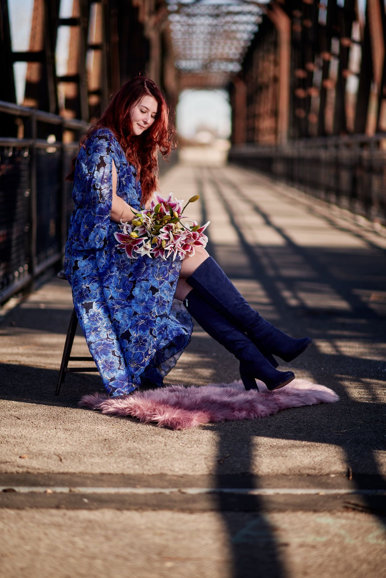 A person in a flowing blue floral dress and boots sits with flowers on a rustic bridge on a sunny day.