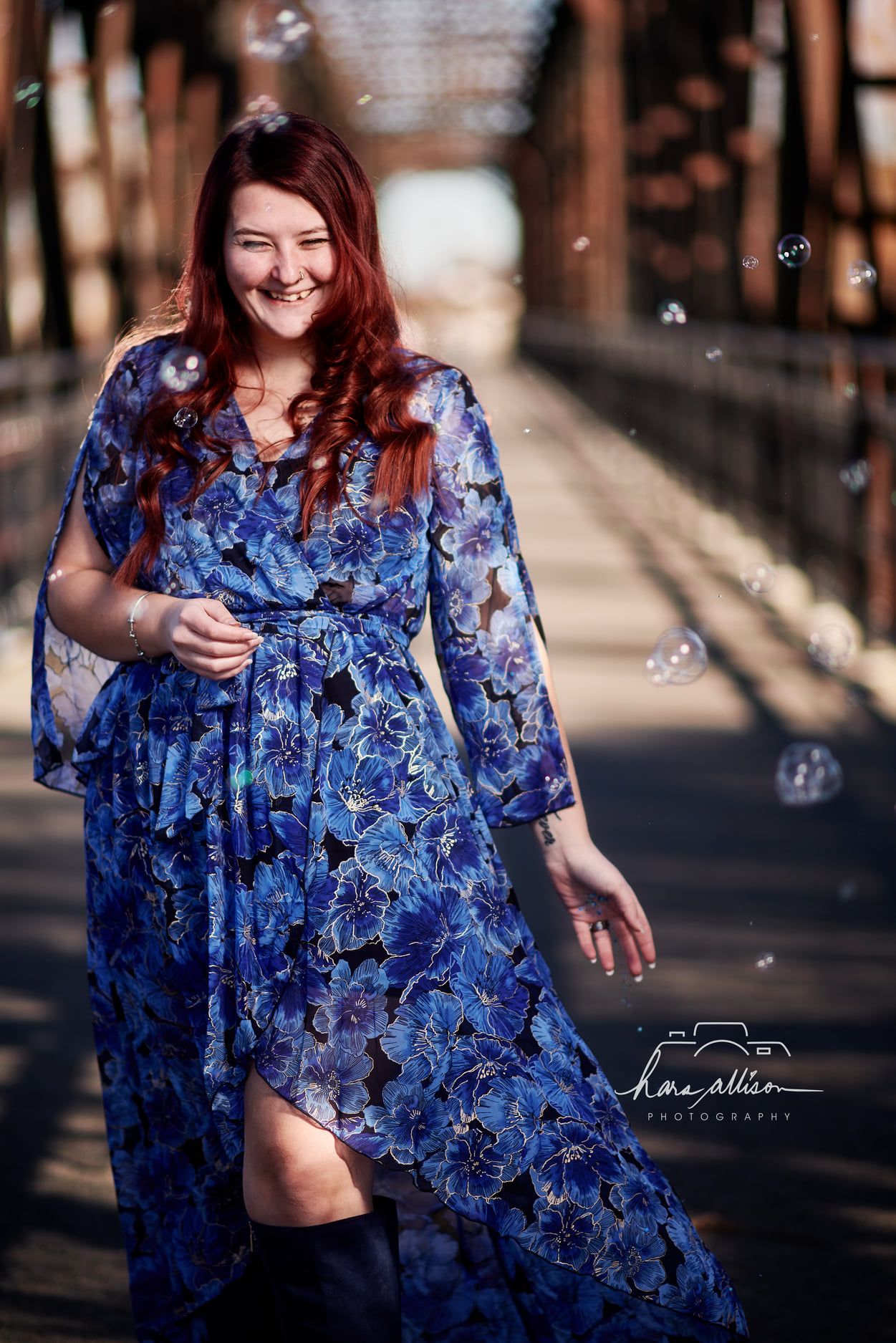 A woman in a flowing blue floral dress poses outdoors with bubbles floating around on a wooden bridge.