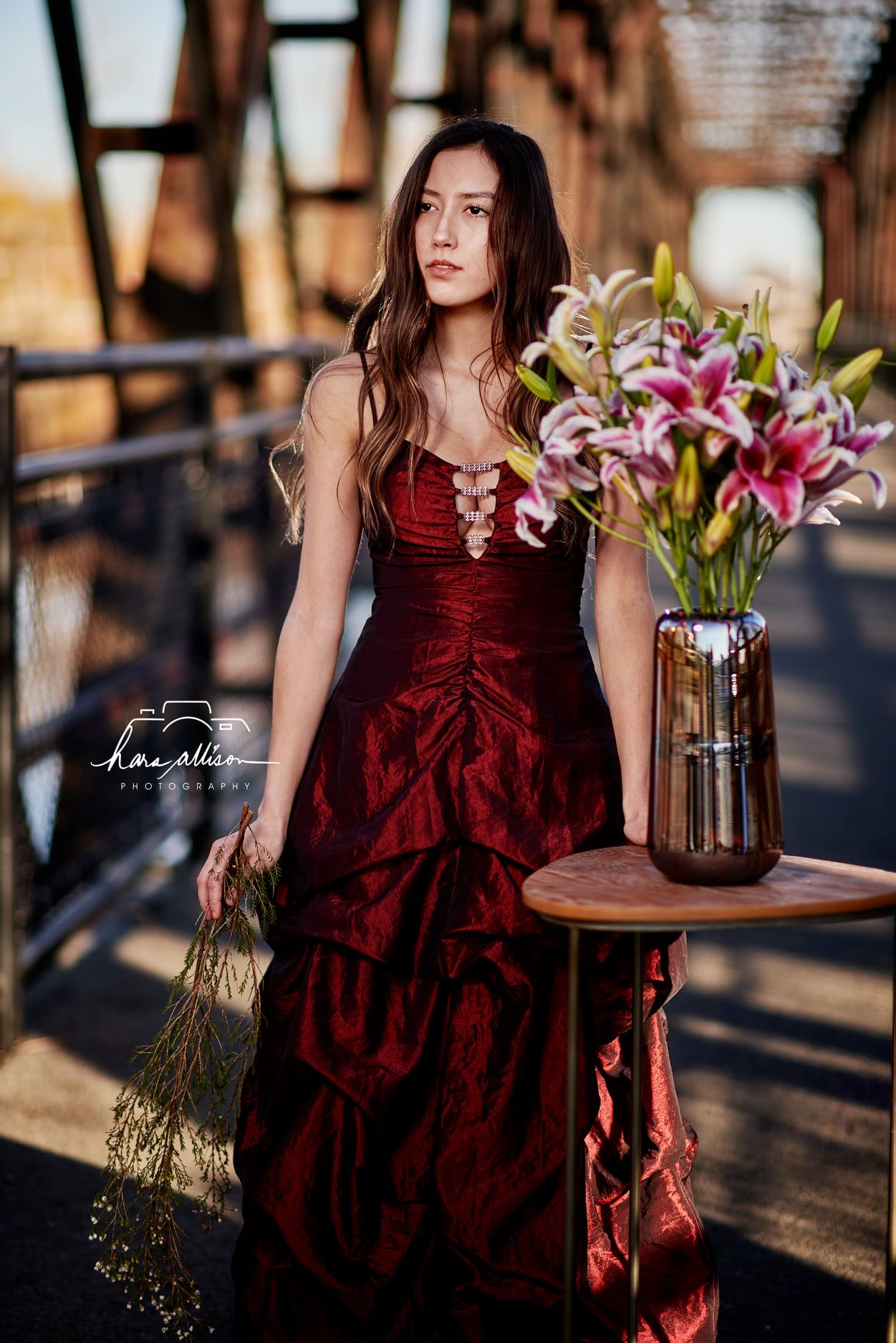 A red velvet gown photoshoot on a bridge with a vase of pink lilies on a wooden table.
