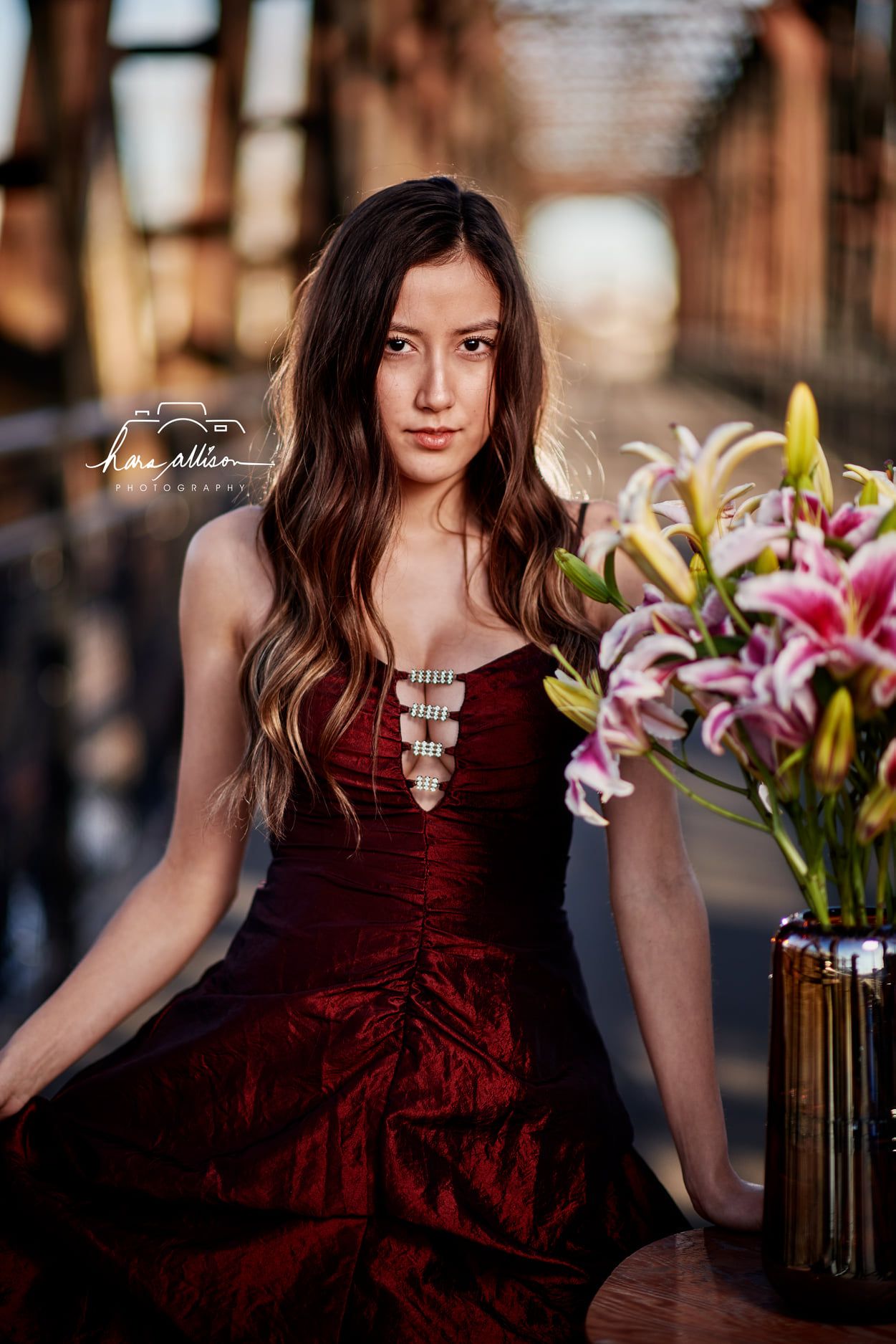 The burgundy velvet dress creates elegant contrast against colorful pink and yellow lilies in an outdoor portrait setting.