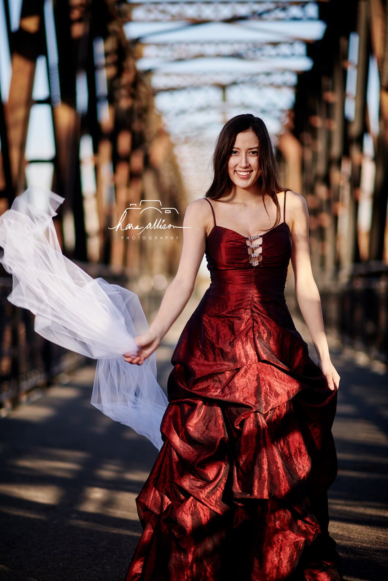 A flowing deep red formal dress photographed in golden sunlight on a rustic bridge with white fabric.