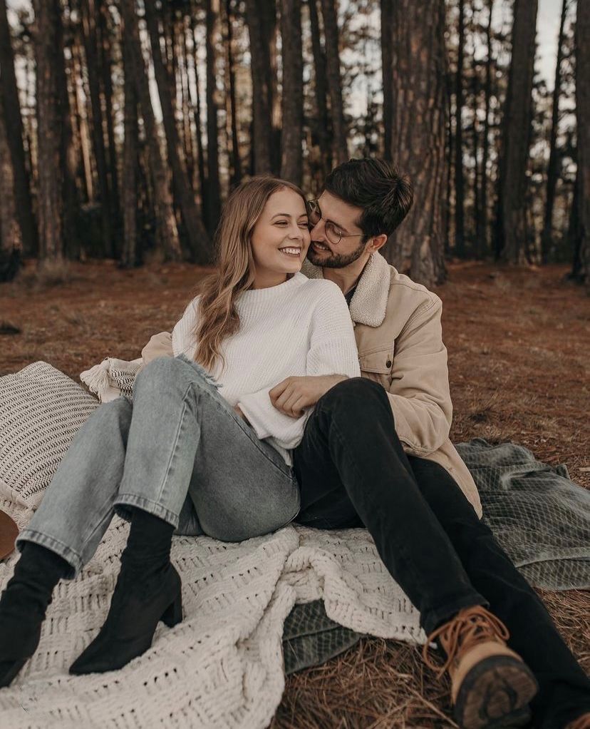 A couple shares an intimate moment while sitting on a blanket in a pine forest during golden hour.