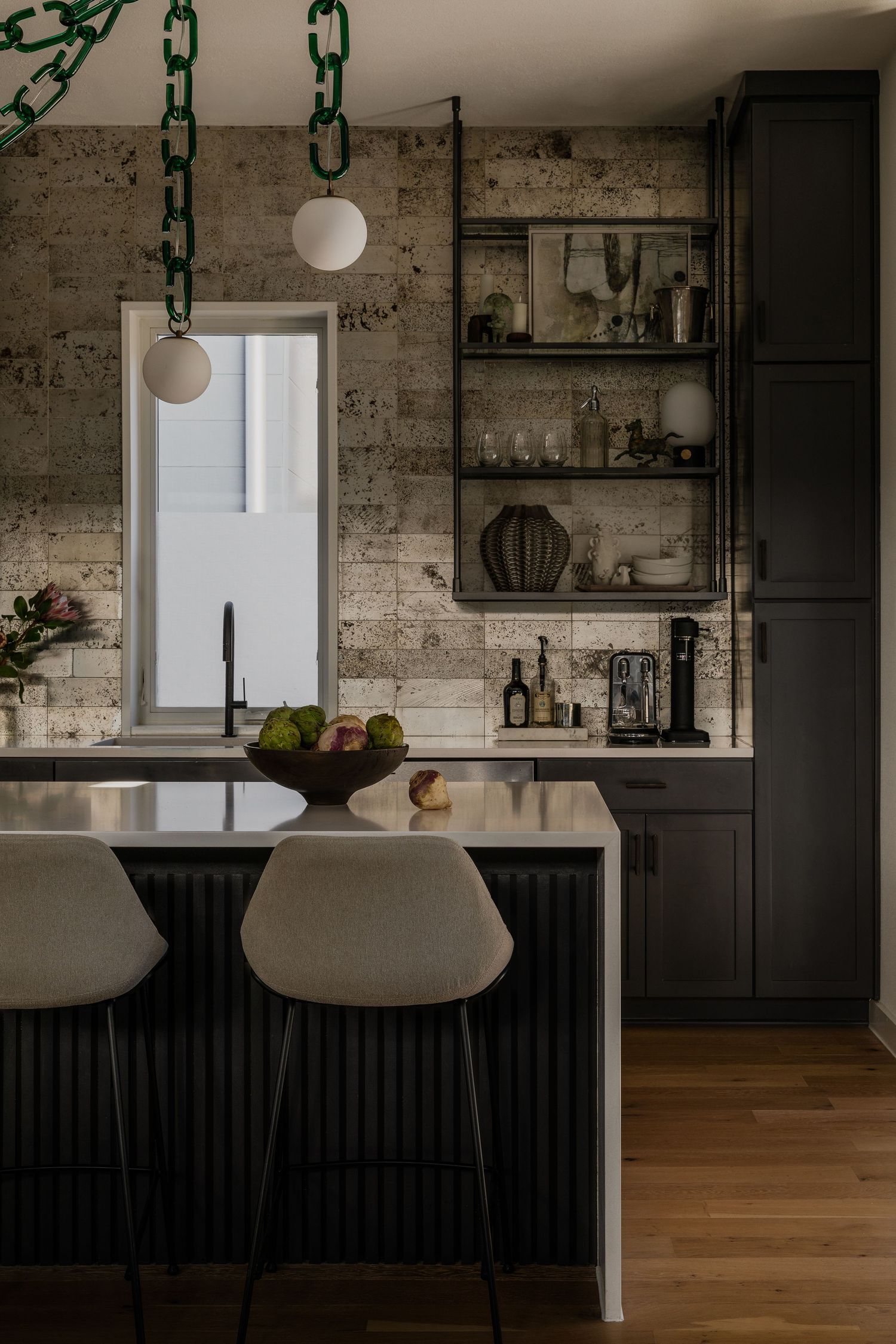 Modern dark kitchen with exposed brick walls, chain pendant lights, cream bar stools and floating shelves displaying glassware.