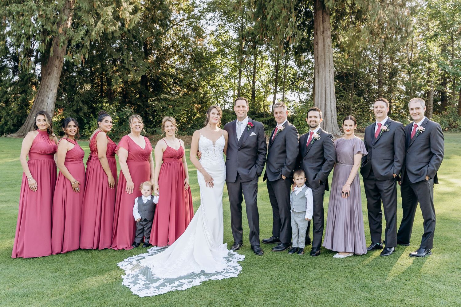 Wedding party in burgundy dresses and dark suits poses together on a lush green lawn.