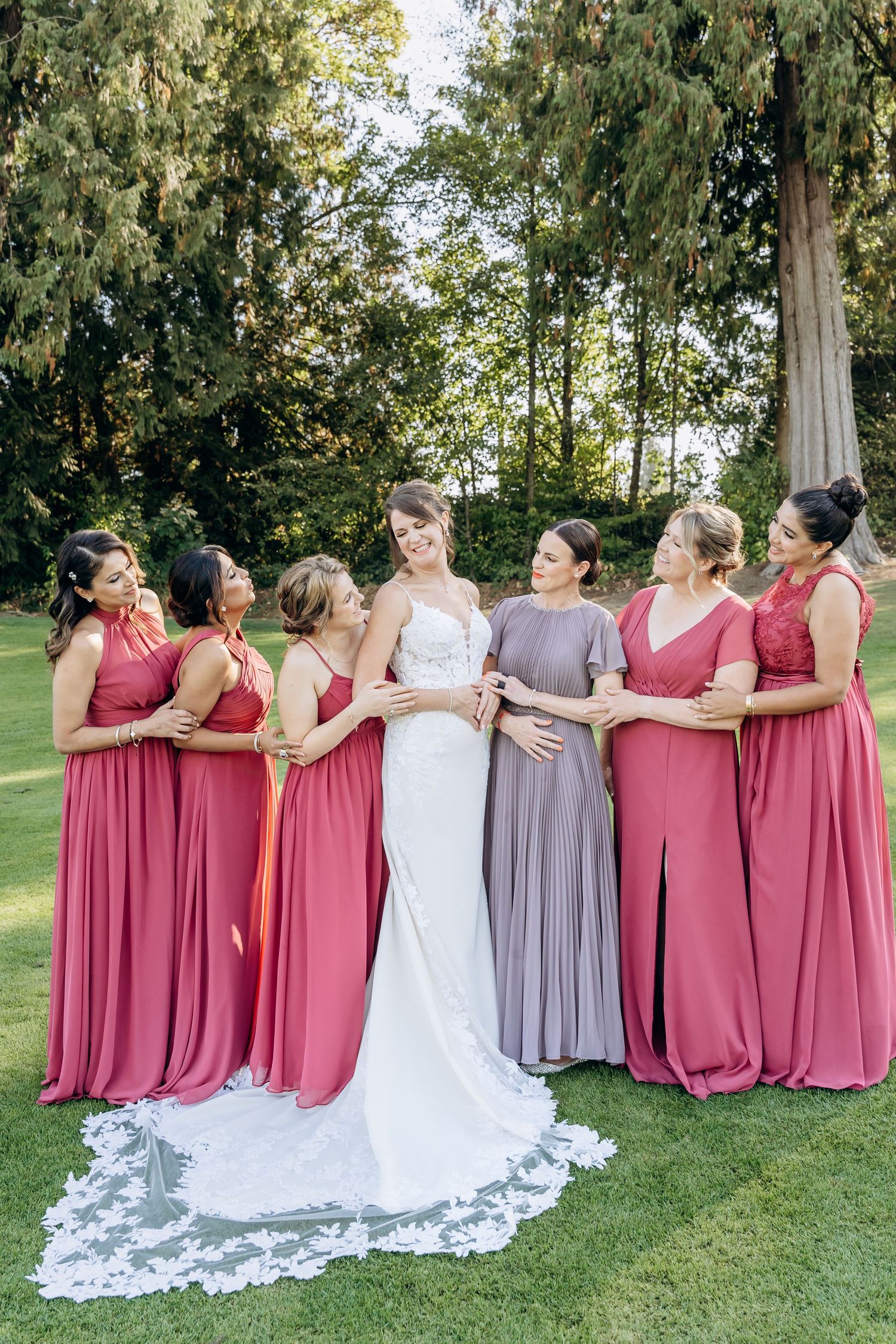 Bridesmaids in rose-colored dresses stand together with bride in white gown outdoors.