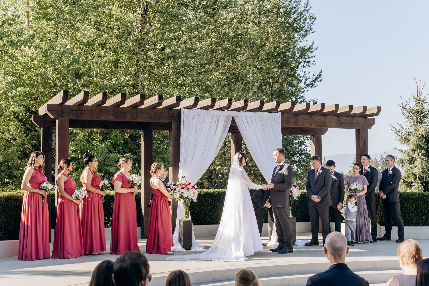 An outdoor wedding ceremony setup with white draping and wooden pergola surrounded by guests.