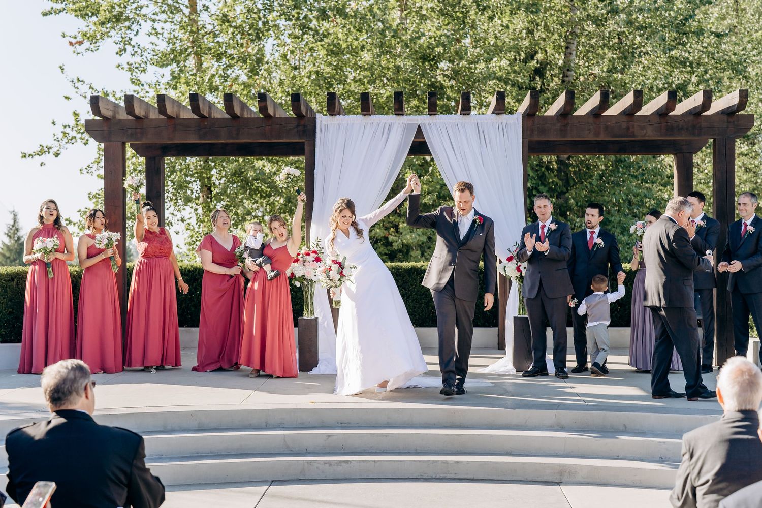 Wedding ceremony site features white draping under wooden pergola with guests seated on both sides.