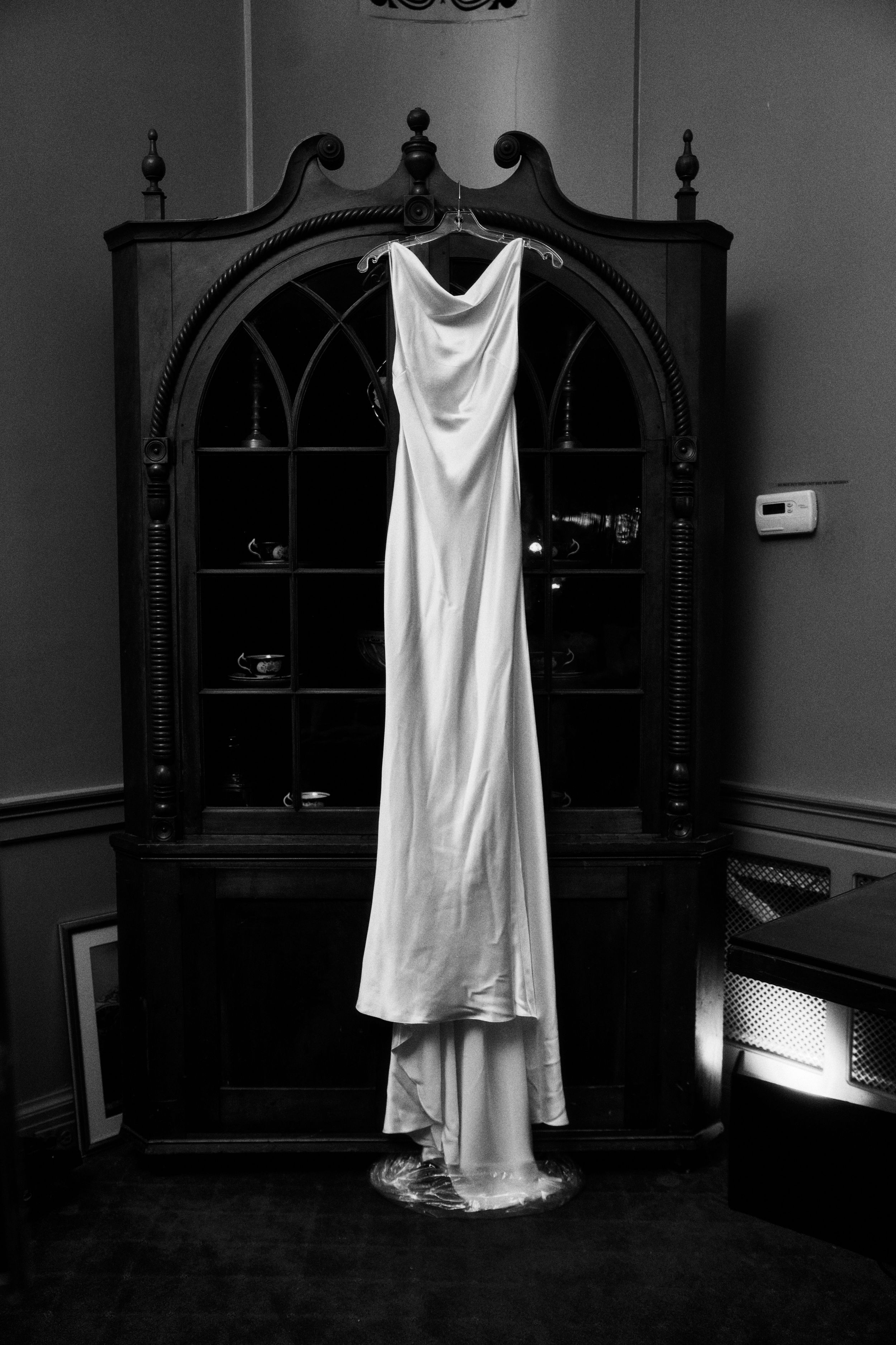 Elegant white wedding dress hanging on ornate black vintage cabinet in black and white.