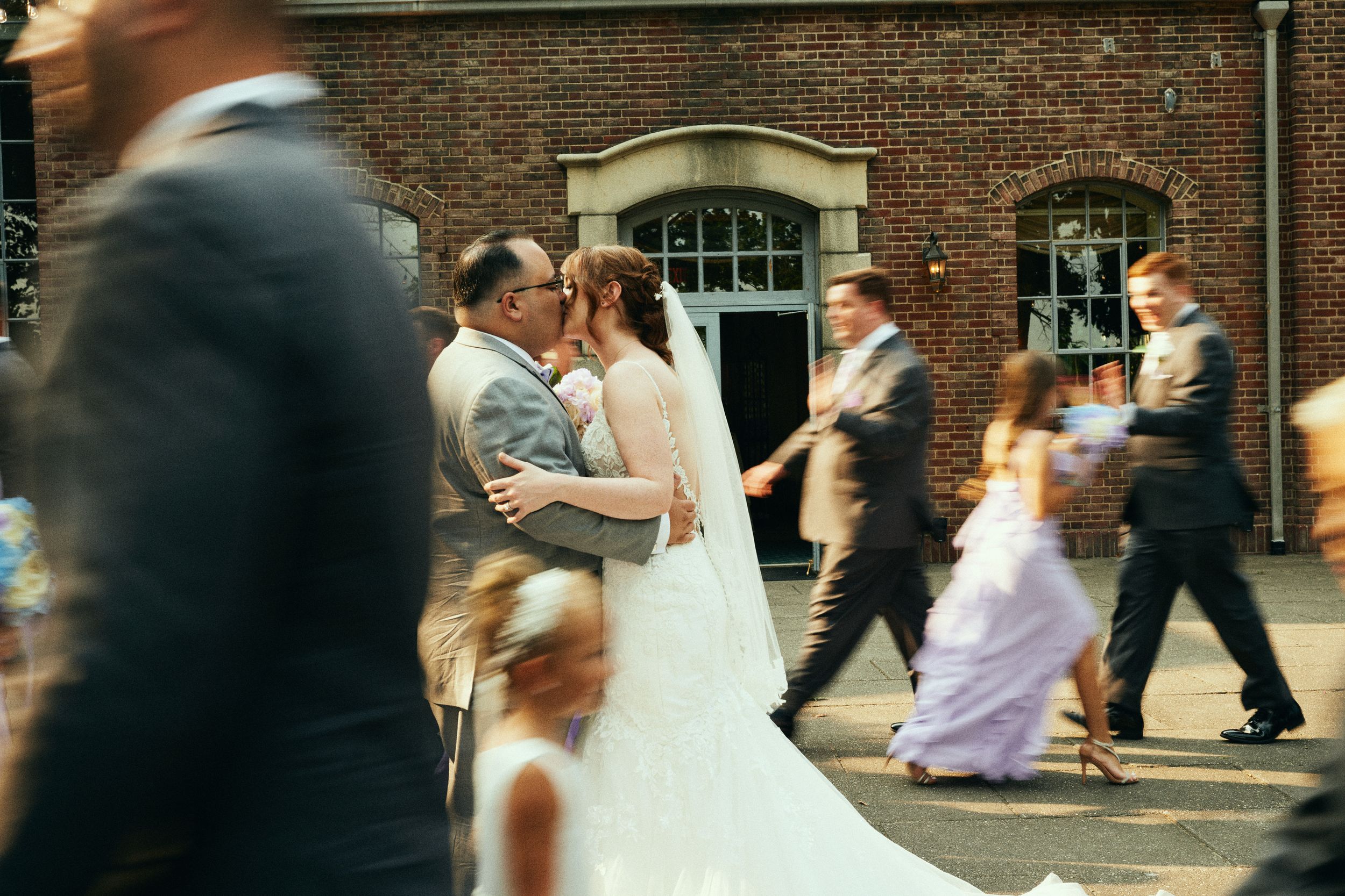 A motion-blurred wedding scene outside a brick building with people walking past the newlyweds.