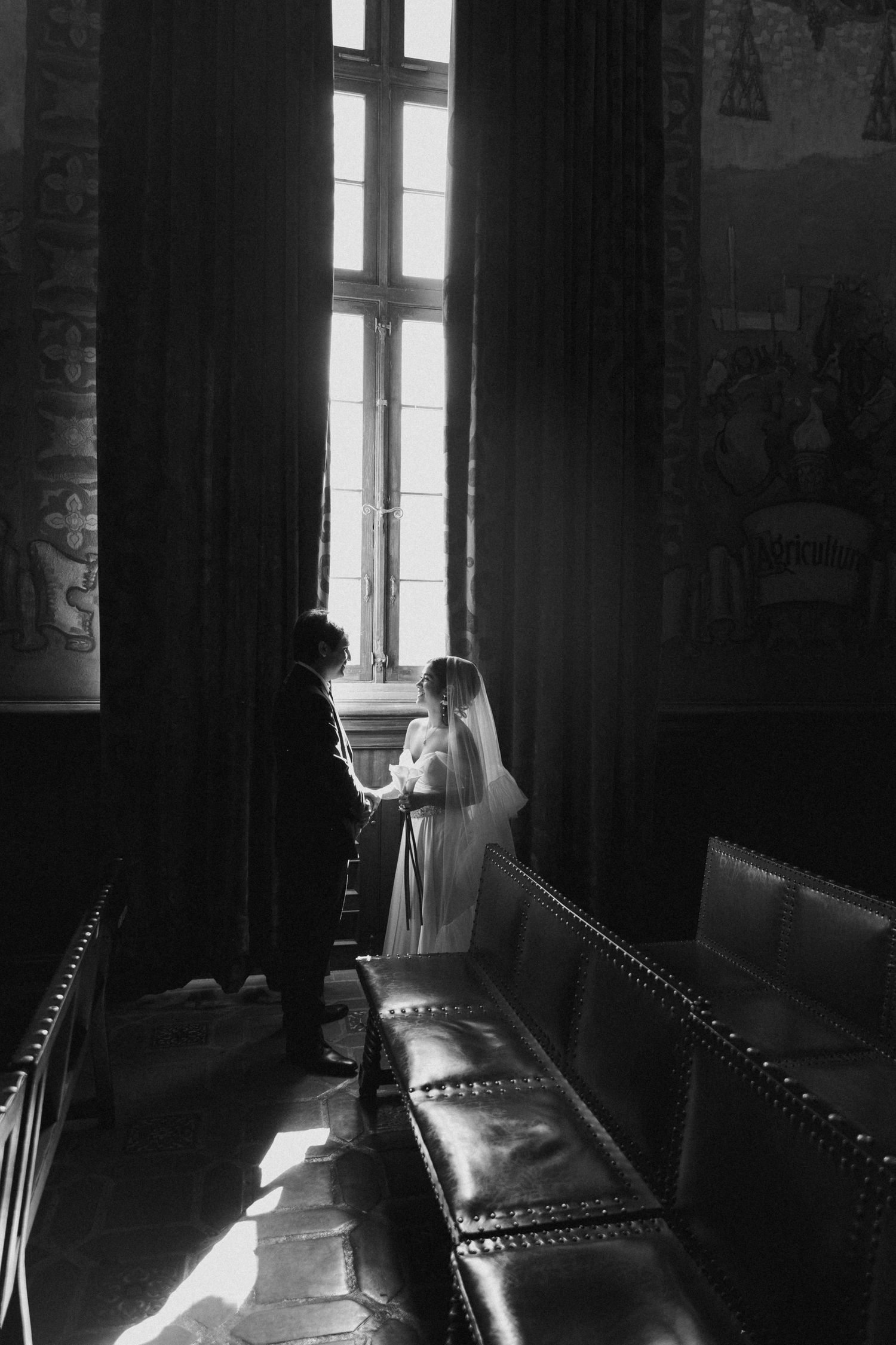 Black and white photograph of bride and groom standing by a tall church window with leather pews in the foreground.