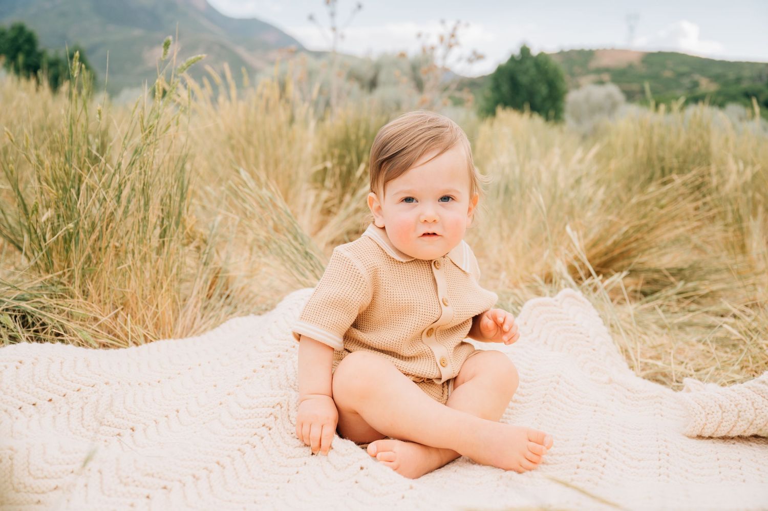 Small baby sitting on a white blanket surrounded by tall wheat grass with mountains in the background.