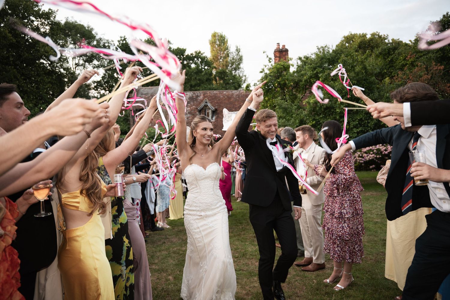 Wedding guests wave pink and white streamers during an outdoor celebration at a garden ceremony.