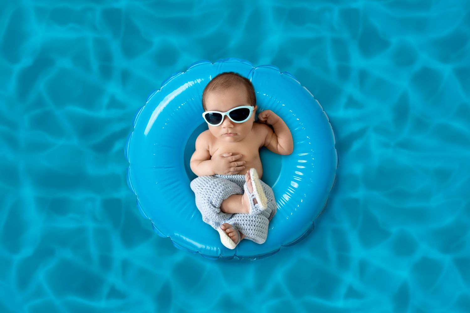 Newborn wearing sunglasses floating on blue pool float in artistic photography setup. Photo by Dani De La Rosa, DigitalMyst Photography