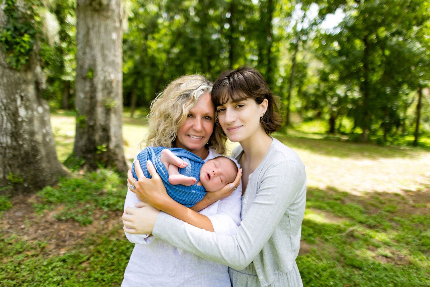 Family members sharing a tender moment while holding a baby outdoors in a sunny garden setting. Photos by DigitalMyst Photography