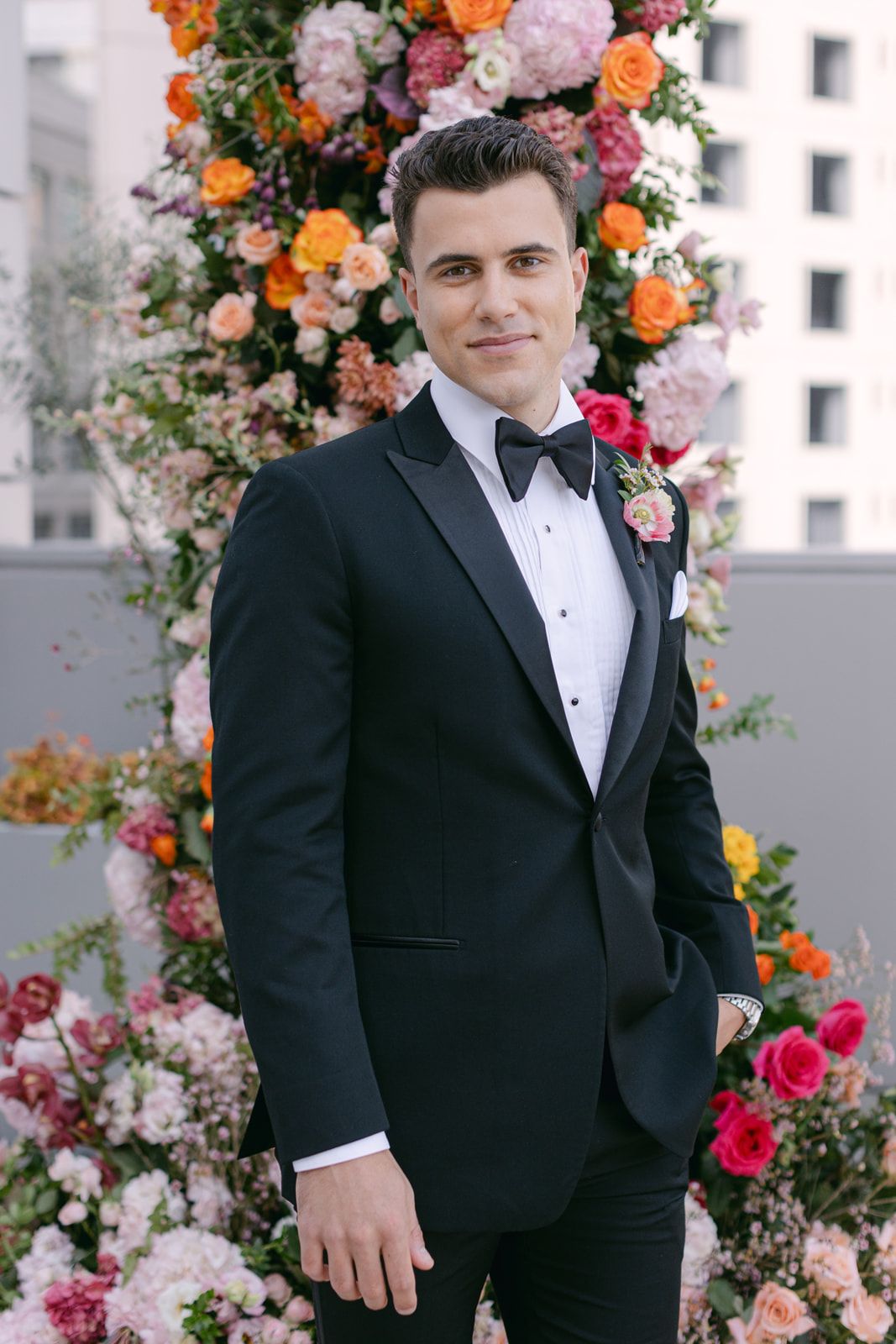 A formal wedding portrait featuring someone in a black tuxedo standing next to vibrant floral arrangements of orange and pink blooms.