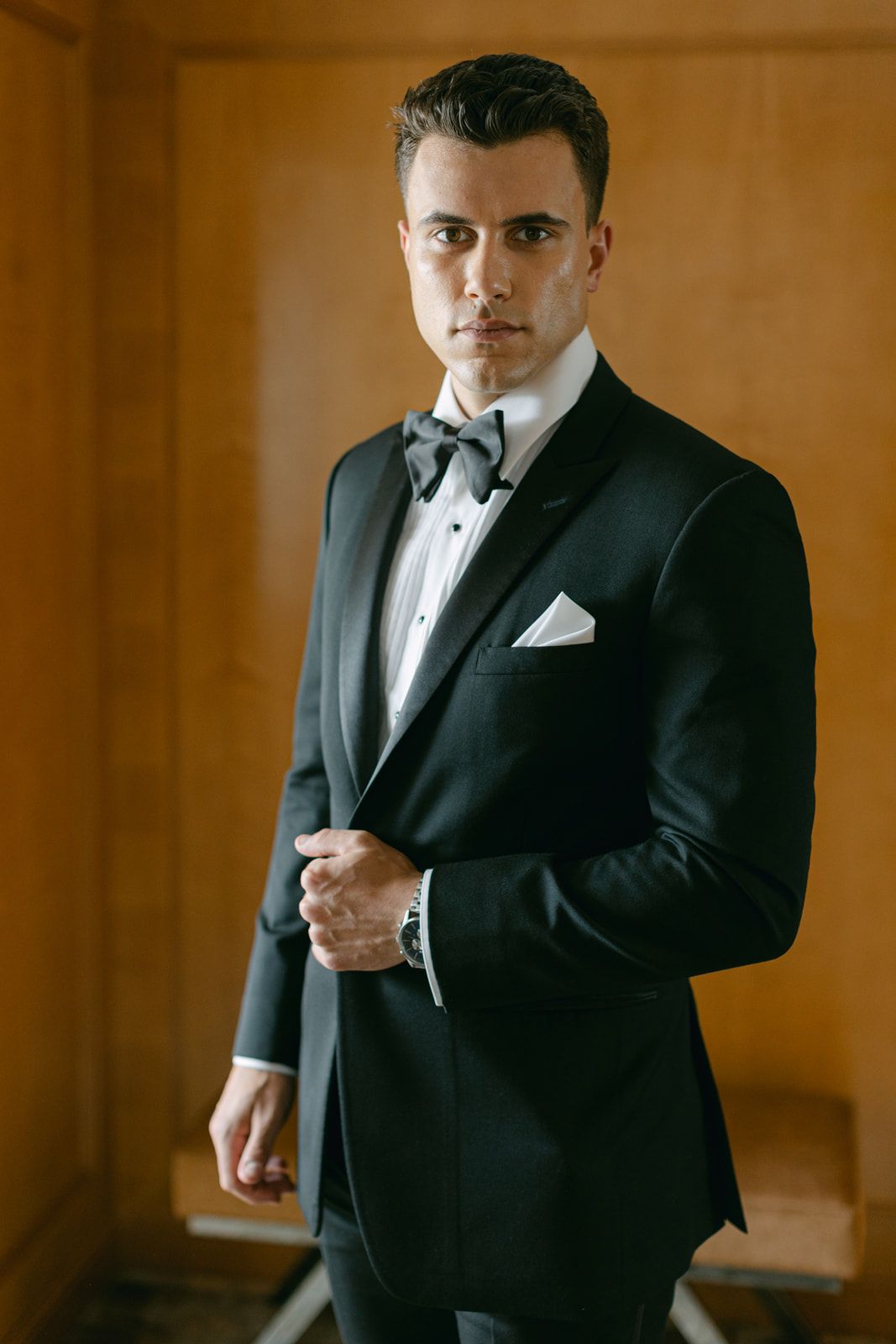 Elegant groom in black tuxedo and bowtie adjusts his watch while getting ready in warm-lit room, shown in multiple poses.