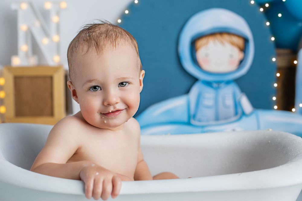 Small child smiling in a bathtub with a blue space-themed wall decoration in the background with twinkle lights. camira cake smash