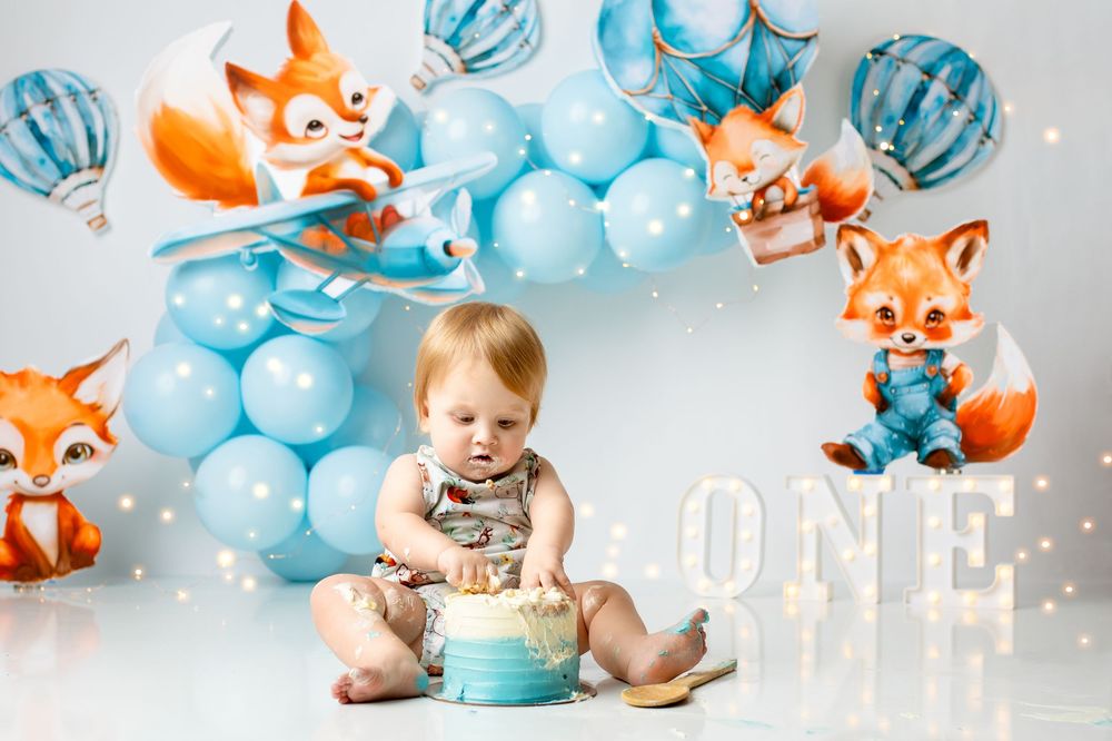 First birthday cake smash setup with blue balloon arch and cute illustrated fox decorations on gray background. raceview cake smash