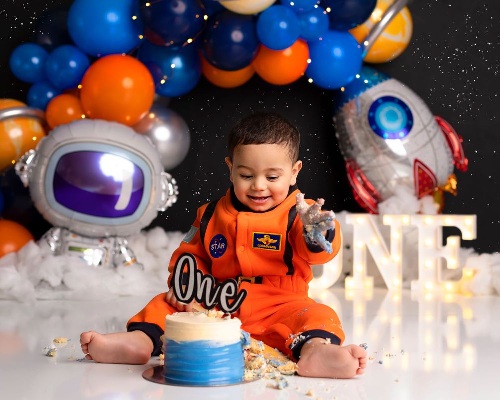 A space-themed first birthday party setup with astronaut balloons, blue cake, and a small child in an orange NASA jumpsuit. pallara cake sm
