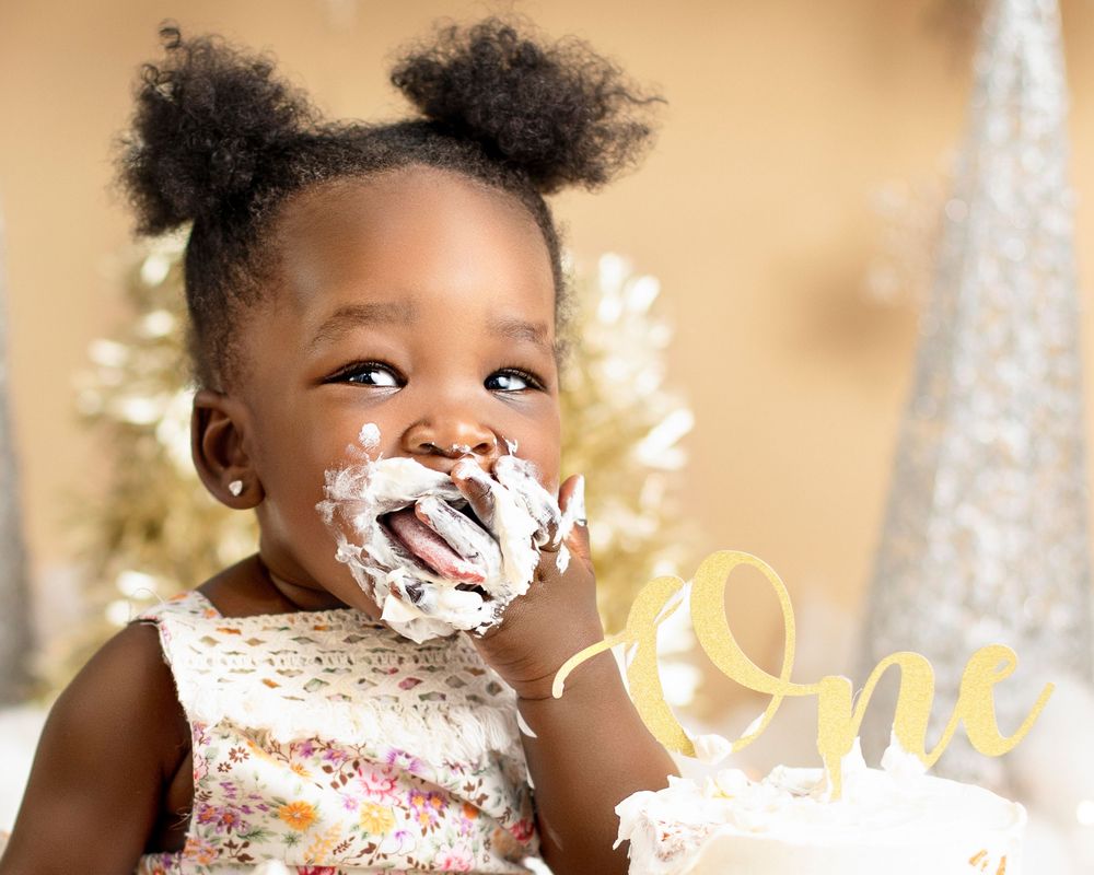 Adorable toddler with puffs hairstyle wearing lace dress smiling with cake frosting all over their face. south sudanse baby first brithday.