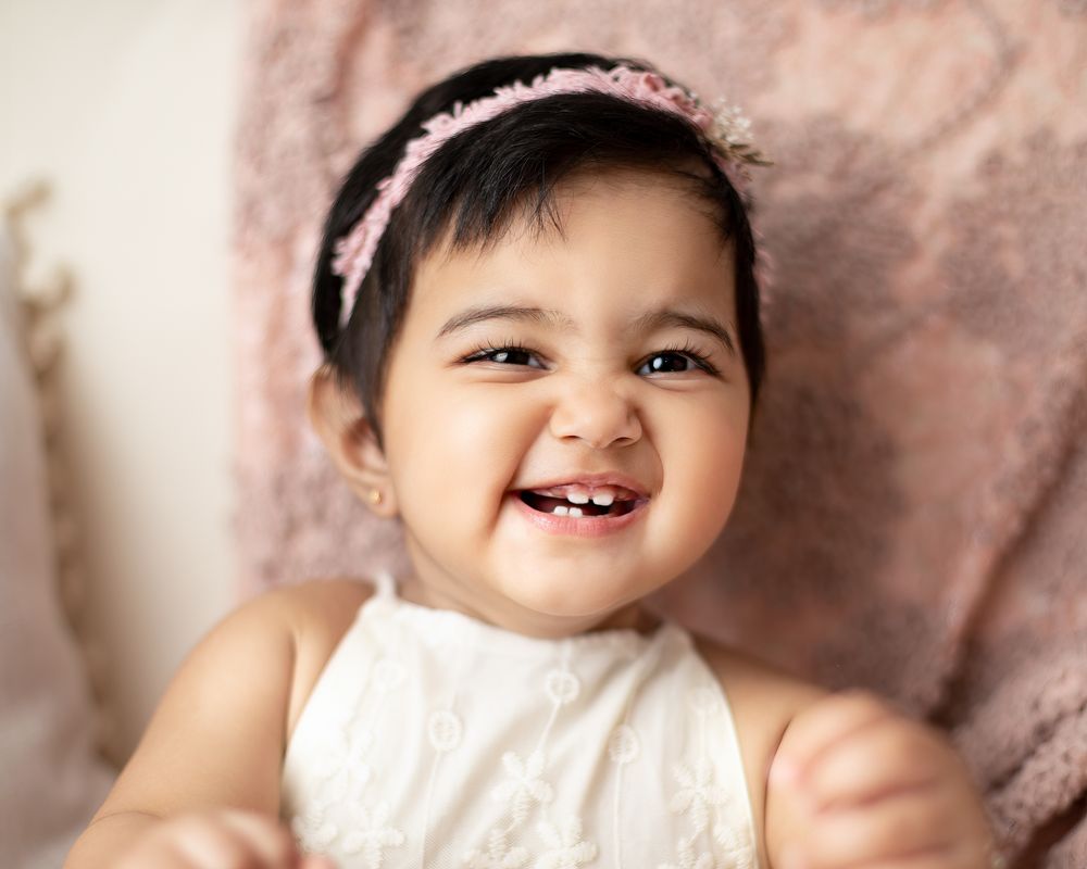 A smiling baby in a white dress and floral headband sits against a soft blush pink backdrop. first birthday, princess theme.