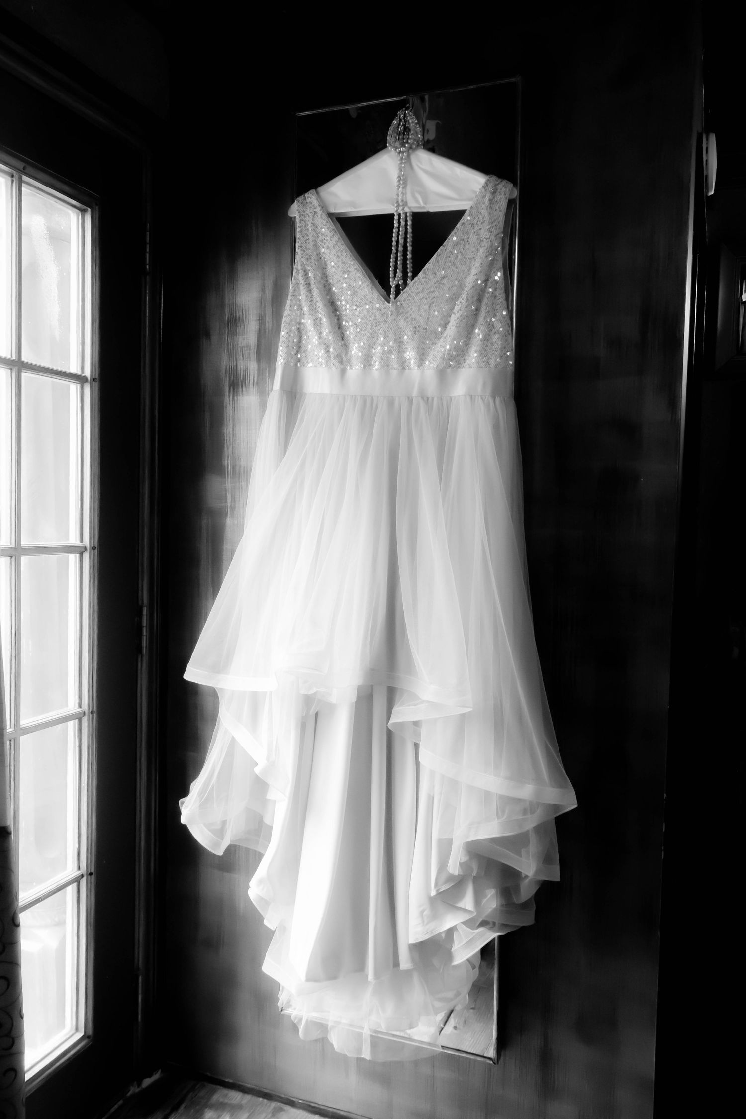 A white wedding dress with lace bodice and flowing chiffon skirt hangs elegantly by a window in black and white.