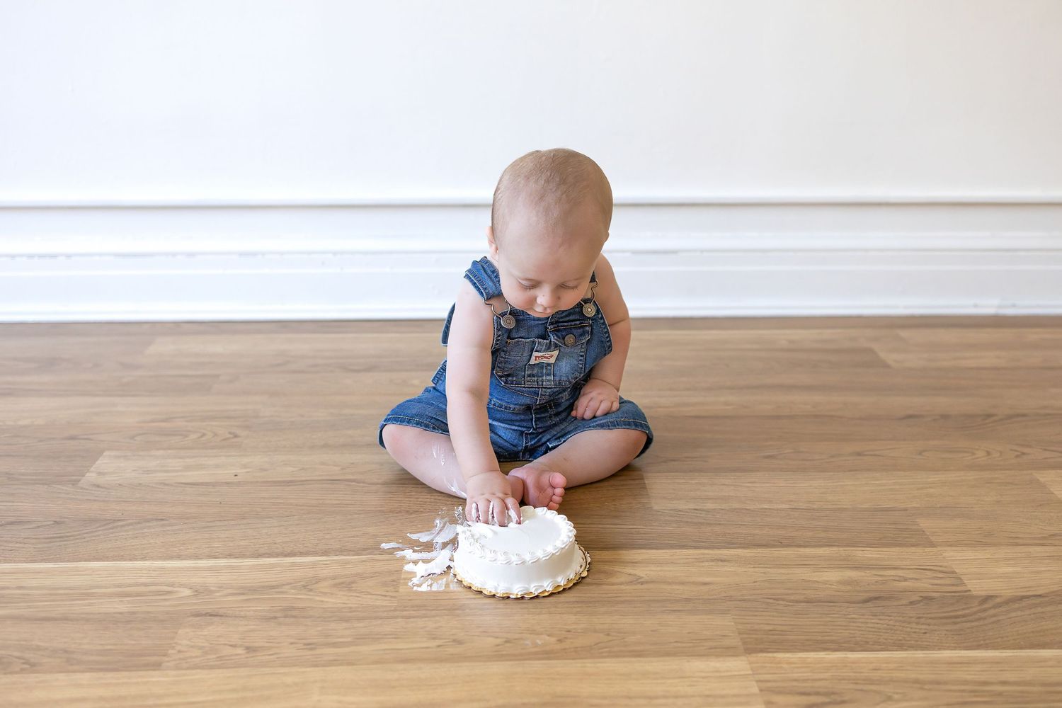 Baby in denim overalls sitting on hardwood floor playing with white frosting or cream.