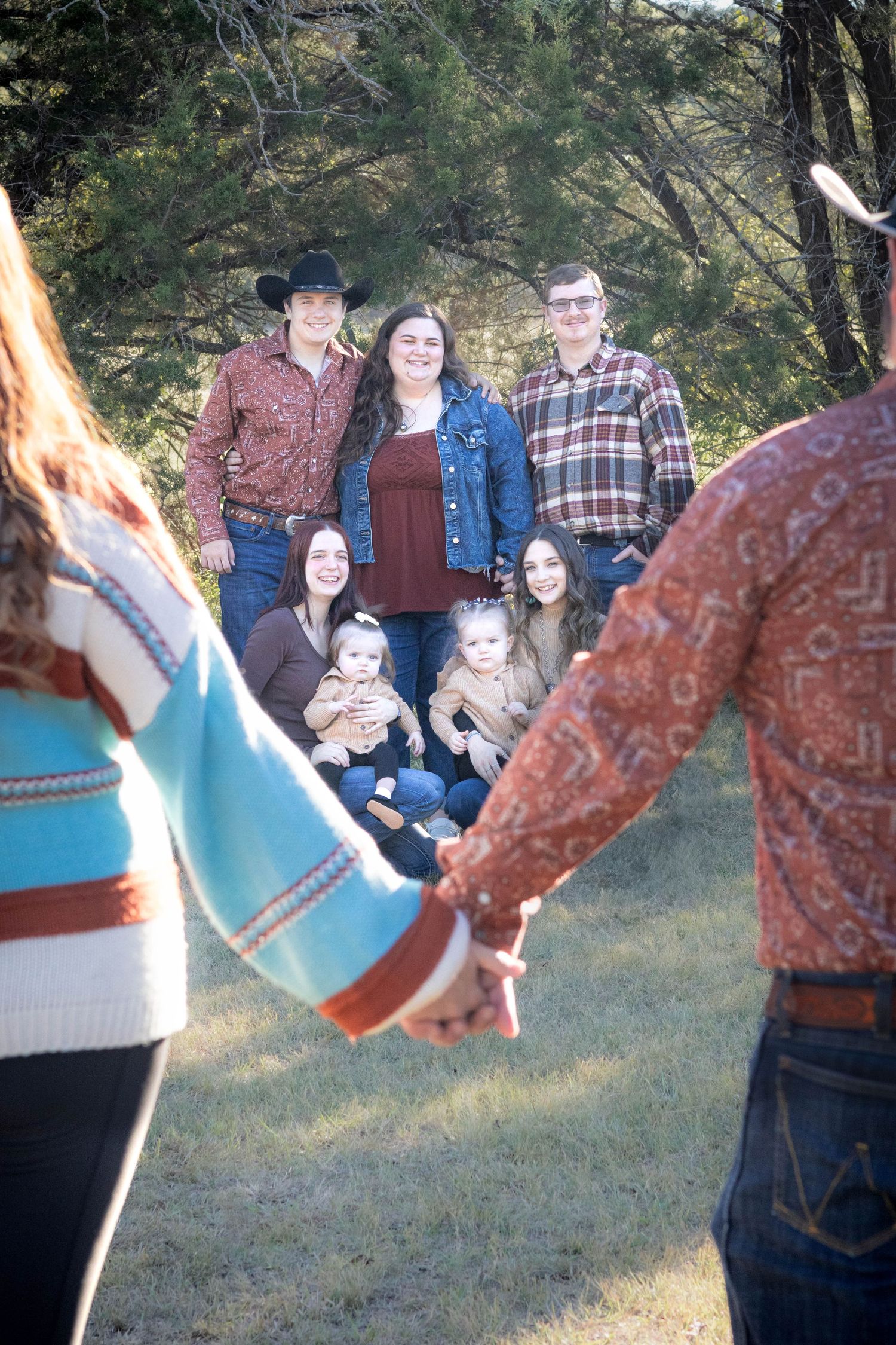 Family group poses together outdoors with pine trees in background while two people hold hands in foreground.