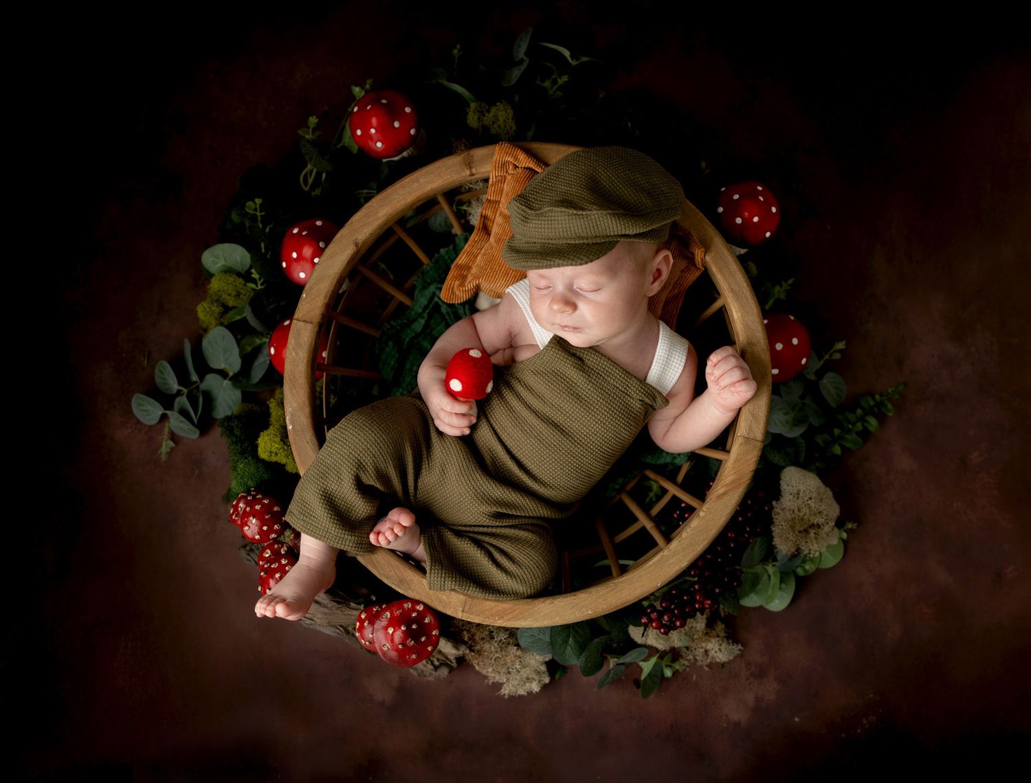 Small sleeping infant in olive wrap nestled in round wooden basket surrounded by red berries and greenery.