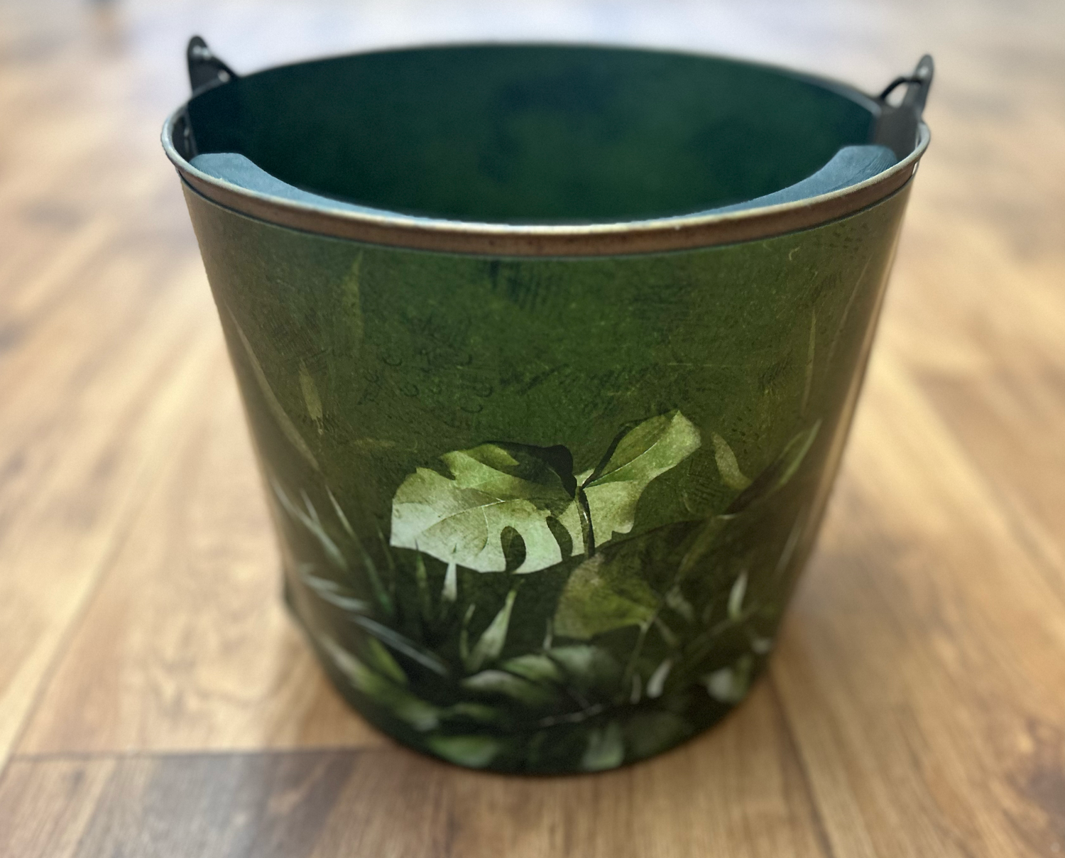 A dark green metal bucket decorated with tropical monstera leaf pattern sits on a wooden surface.