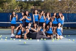 Tennis team members sit together on blue court during practice session in outdoor setting.