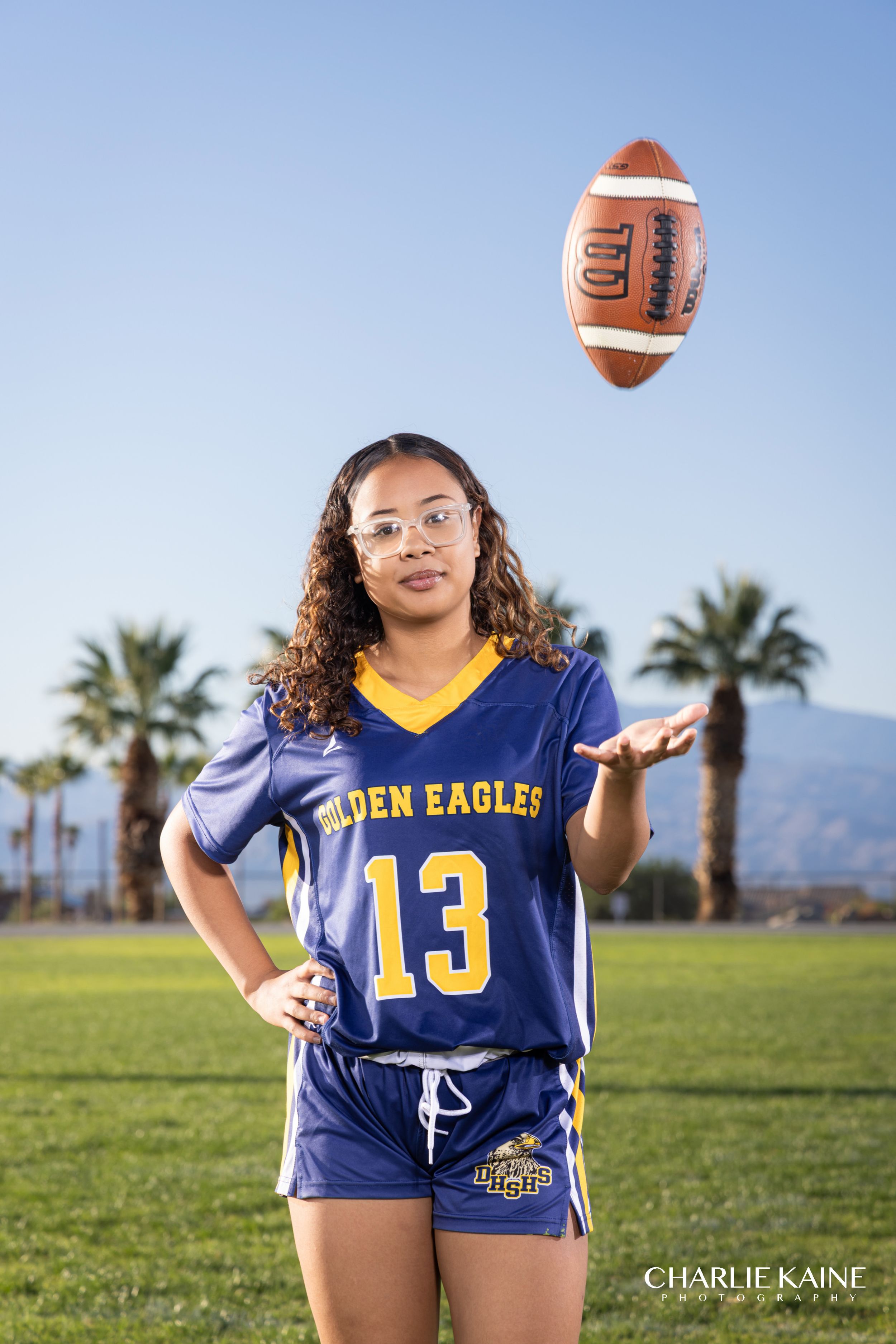 Football player wearing blue and gold uniform number 13 poses on sunny field with palm trees in background.