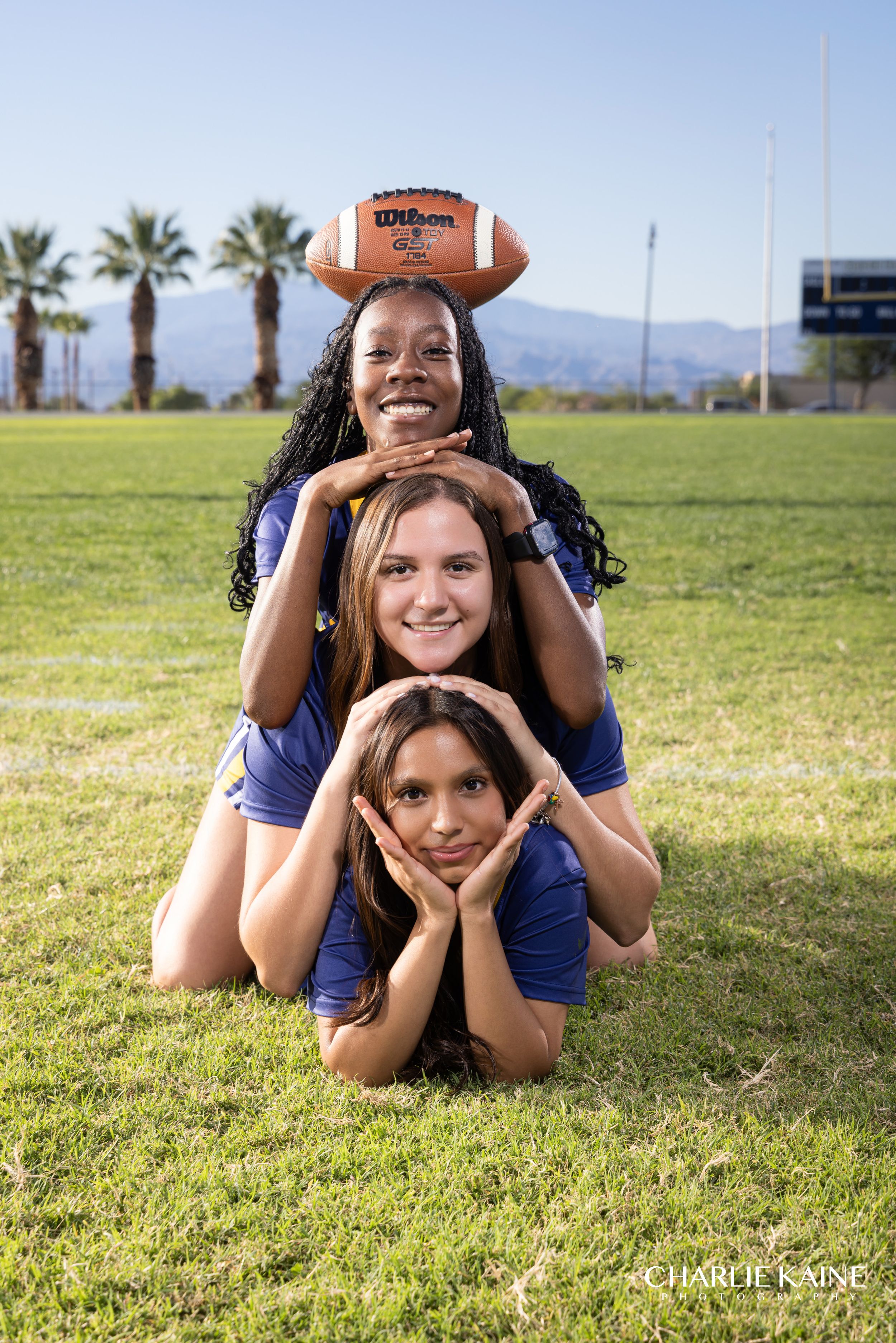 Three teammates in navy blue jerseys pose playfully with a football on a sunny grass field with palm trees in background.