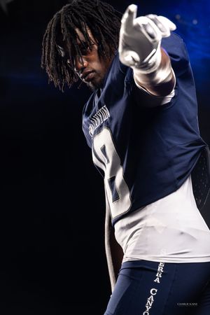 Football player in navy uniform pointing forward dramatically against dark background.