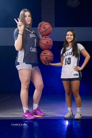 Two basketball players pose with basketballs in navy and white uniforms on a dark court with dramatic lighting.