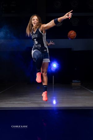 Basketball player jumps in mid-air while dribbling on dark court with dramatic blue lighting effects.