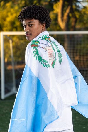 Athletic figure wearing light blue and white flag or cape on sports field at sunset.