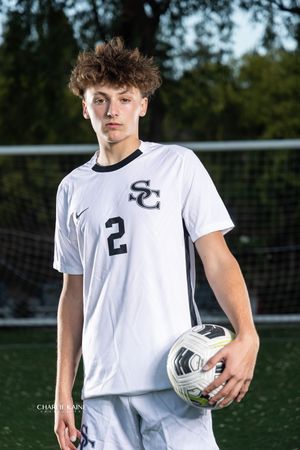 Soccer player in white uniform holds ball on field with goal nets visible in background.