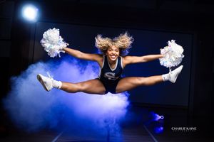Cheerleader performs aerial split jump with pom-poms amid dramatic blue smoke effects.