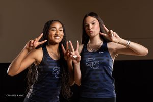 Two athletes in matching navy tanks make peace signs against brown background.