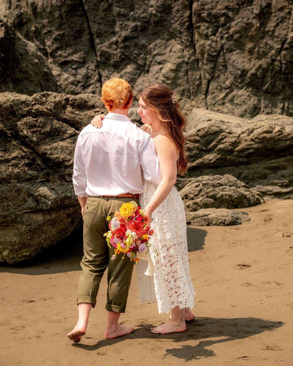 A couple walking along a beach with rocky cliffs and colorful flowers.