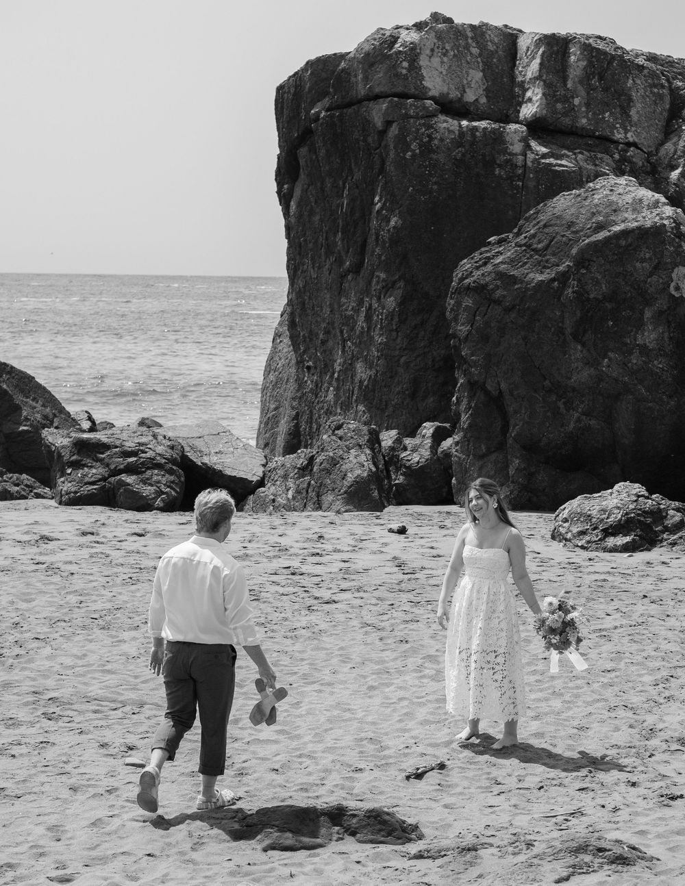 A black and white photo of two people walking near large coastal rock formations.