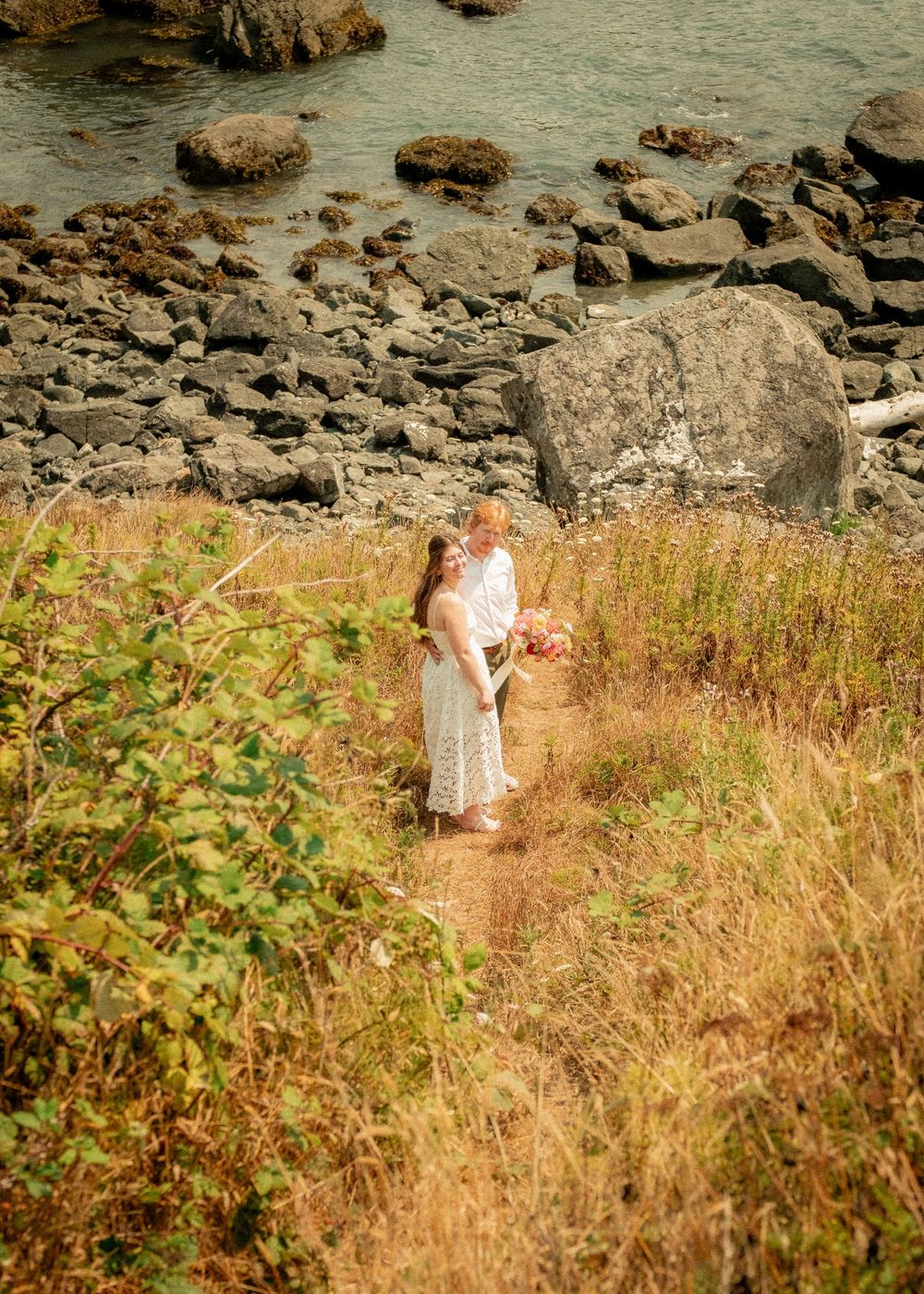A couple walking through tall grass near a rocky beach landscape.