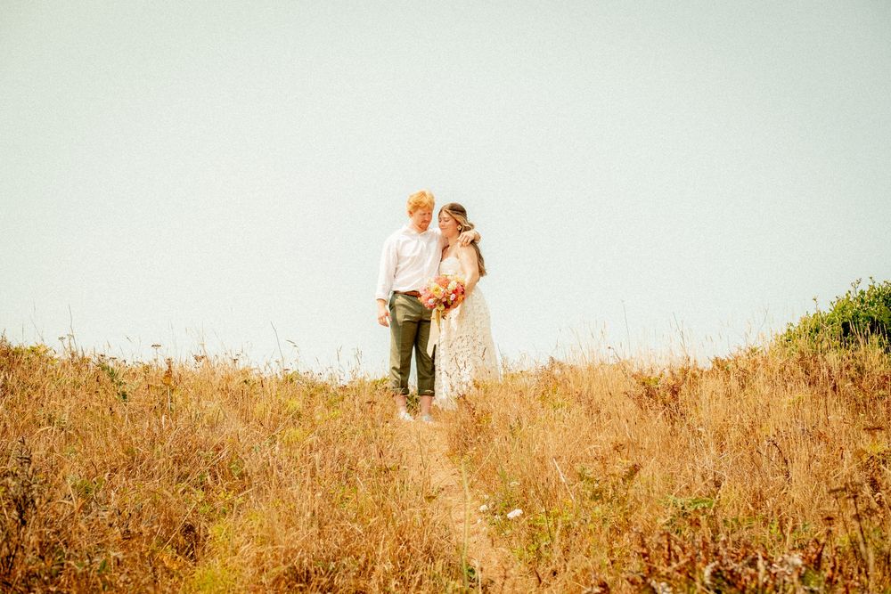 Two figures standing in a sunlit field of tall golden grass near the ocean.