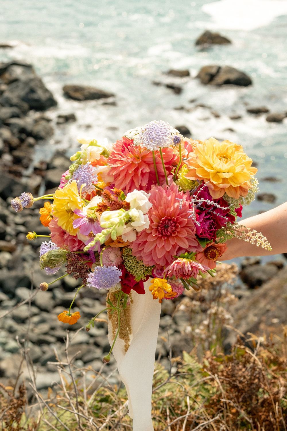 A vibrant bouquet of dahlias and wildflowers against a coastal ocean backdrop.