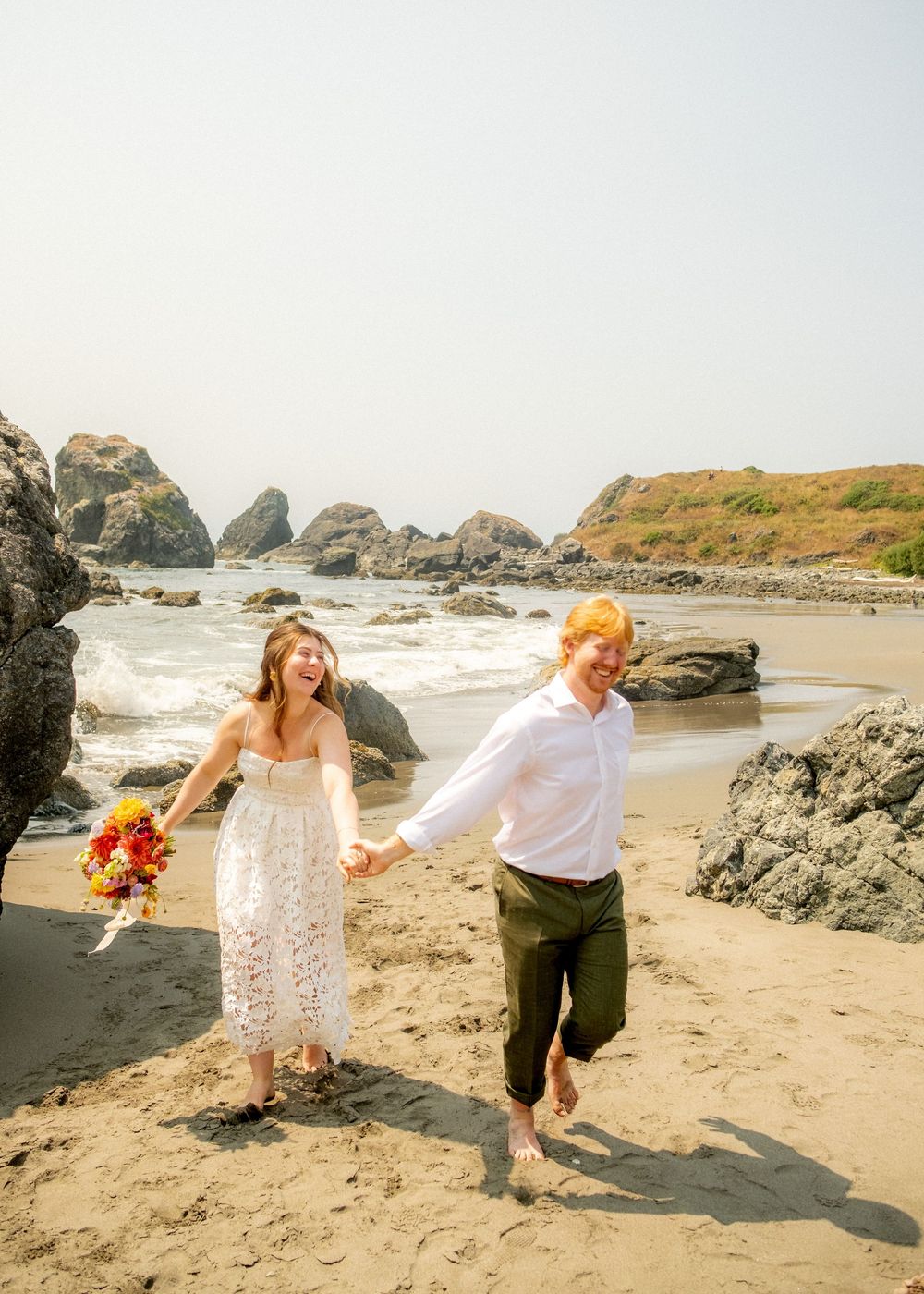 A couple runs joyfully on a beach with large rock formations and crashing waves in the background.